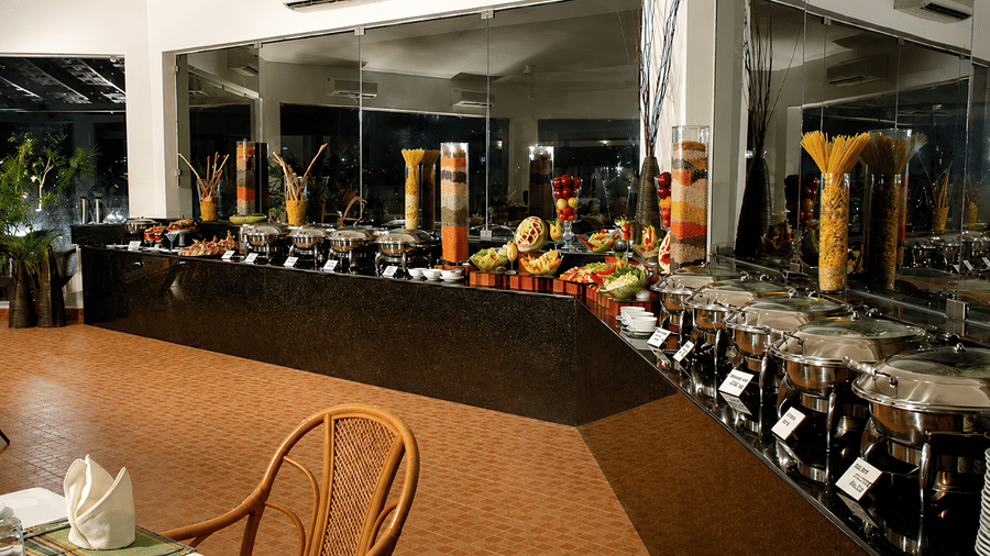 Restaurant buffet counter with colorful juice dispensers and wicker chairs at Chariot Beach Resort, offering a bright and relaxed setting.