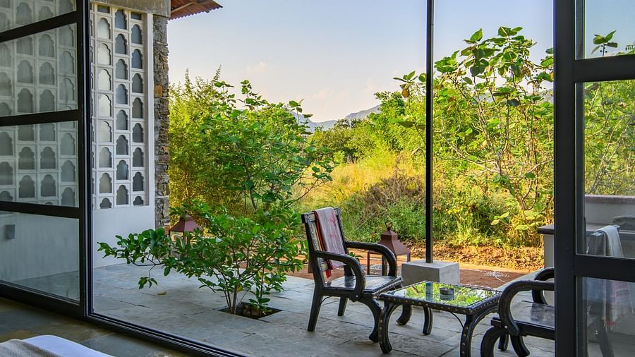An image of a room with a queen size bed, chandelier, ceiling fan, other decorative items and an expansive glass door leading out - Utsav Camp Sariska