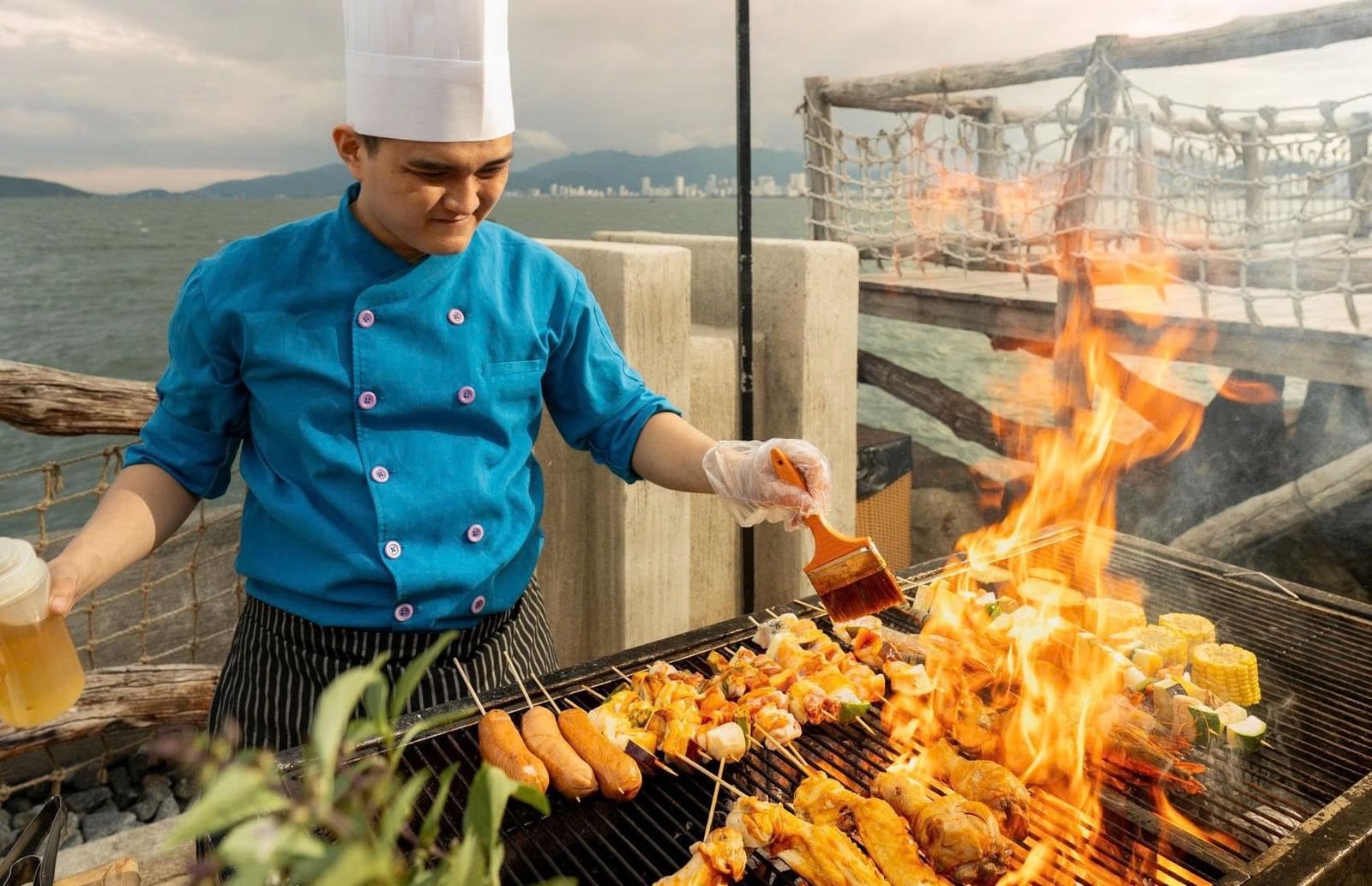A chef in a tall white hat and a blue uniform is grilling food on a barbecue at Alibu Resort Nha Trang.