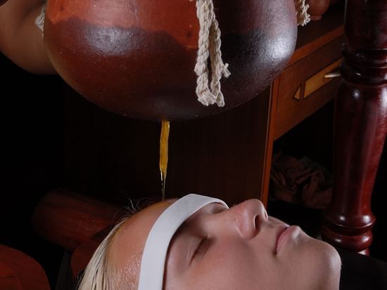 Woman experiencing Shirodhara therapy as warm oil gently streams onto her forehead from a traditional clay pot.