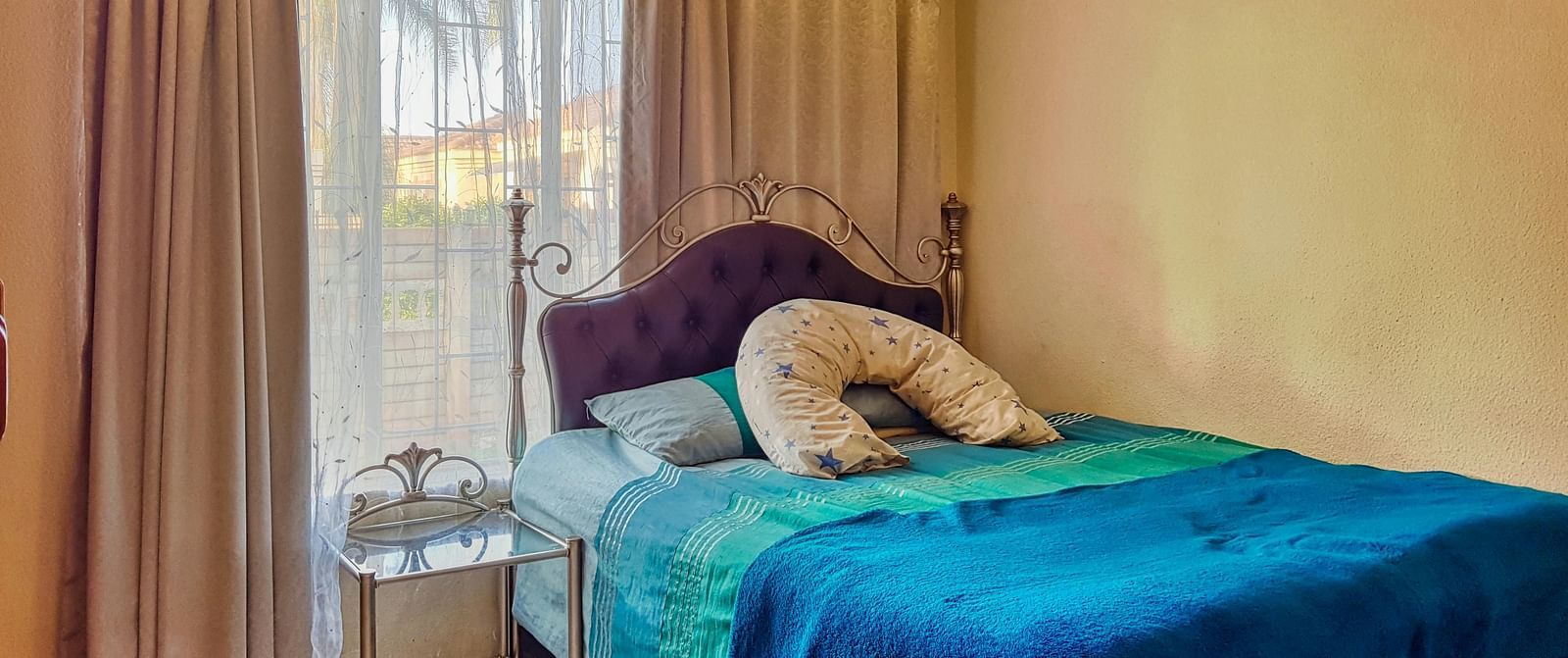 A bedroom featuring a double bed with blue sheets spread neatly over it with beige curtains covering the window behind it.