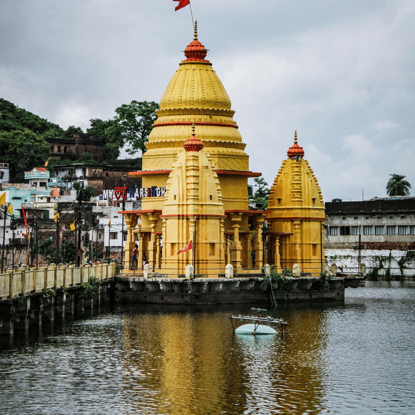 Stone Shiva temple with a tiered spire (shikhara) and a green flag, located near a body of water.