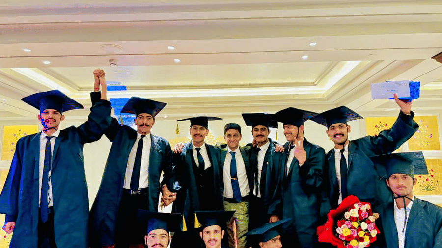 A group of graduates stands together indoors at Clarks Group of Hotels. They wear graduation gowns and caps while holding a bouquet and a box during the celebration.
