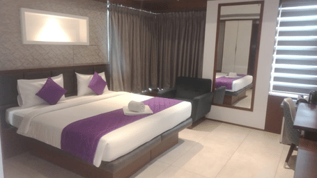Full view of the Executive Suite at SRS Suites, Koramangala, featuring a double bed along with purple-coloured bed runner spread over it with a full-length mirror attached to the wall beside it.