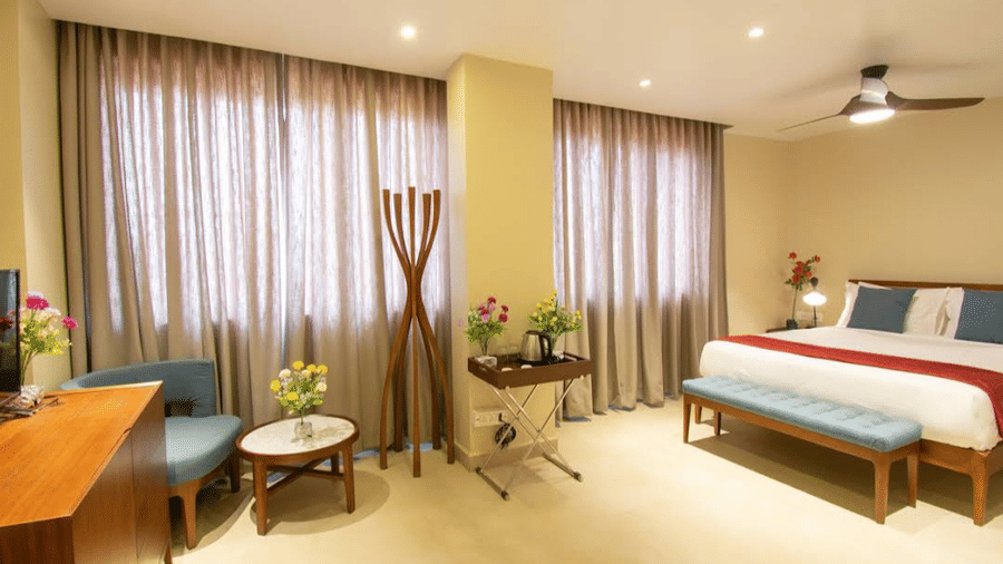 Bright room with a bed, blue chair, windows covered with curtains, and small tables at Estherea Elite, Morjim.