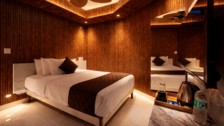 Interior view of the Executive Comfort room at Shakuntalam Hotels featuring a king bed with dark accent pillows rich wooden wall panels ceiling fan and warm ambient floor lighting throughout.