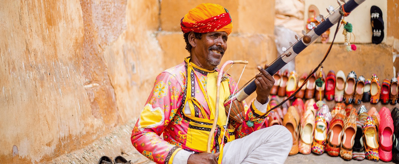 A musician in colourful traditional attire sits on the ground playing a stringed folk instrument, with rows of vibrant handcrafted shoes displayed behind him against an earthy wall.