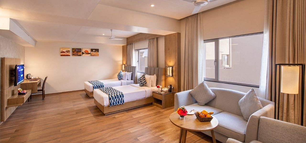 A spacious hotel suite featuring two single beds with blue and white accents, a comfortable seating area with a sofa and armchairs around a small table with fruit, a work desk with a chair, and a television, all on a wooden floor at Lords Inn Motikhavdi