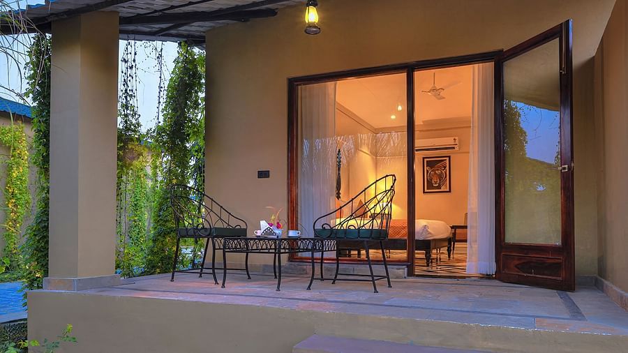 A ground-level view of Superior Cottage's patio featuring a small table and two wrought-iron chairs, with the interior of the room visible through open glass doors - Singhvilas, Ranthambore