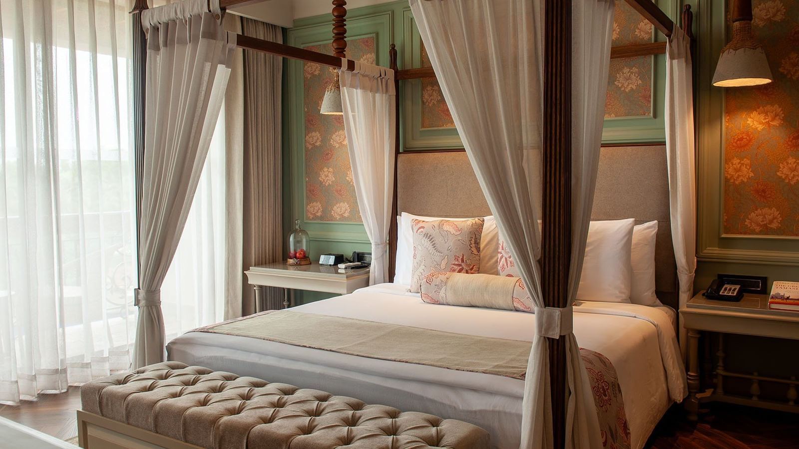  A luxurious suite room at The Evren, Vagator, with a four-poster bed draped in white curtains. The room features a decorative headboard wall with floral wallpaper and a tufted bench at the foot of the bed.