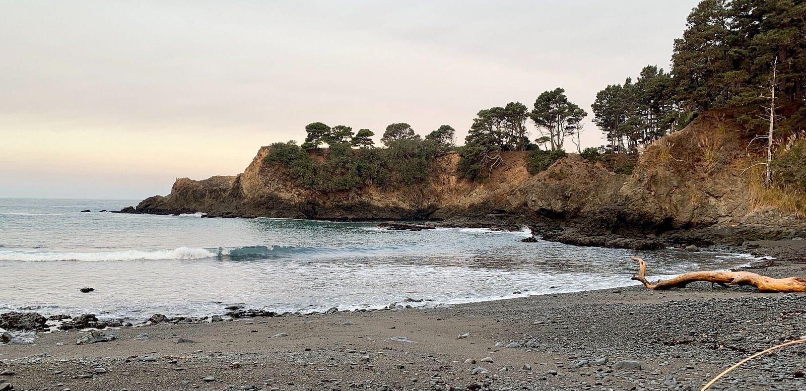 A secluded beach with dark sand and driftwood logs sits nestled between rocky cliffs under a cloudy sky.