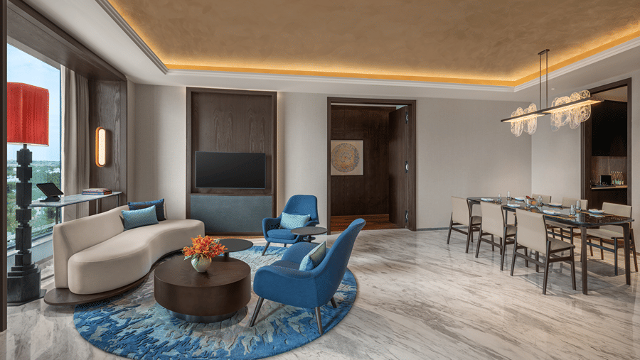 Presidential Suite living room with a curved sofa, blue armchairs on a round rug, a TV, a work desk and a long dining table at The Residency Signature Madurai.