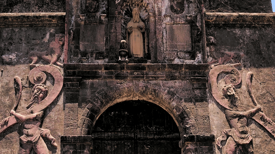 The entrance of an old stone fort, featuring intricate carvings and a cross at the top of the structure, set against a clear blue sky.