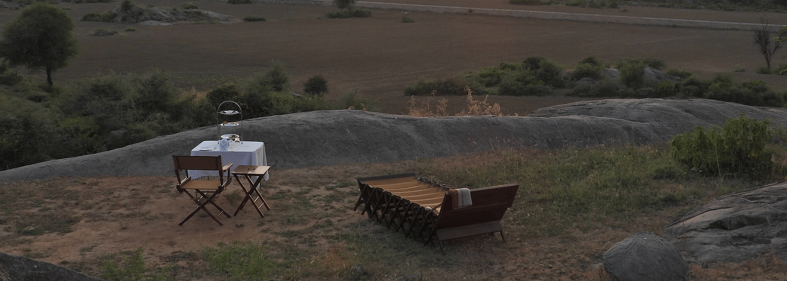 An image of camping site where a chair and table is placed near a lounger bed, and the sun is also setting at the horizon - Utsav Camp Sariska