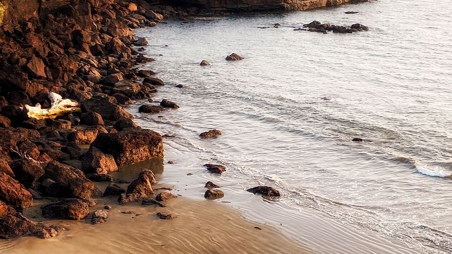 An overhead view of a small, secluded beach with dark rocks and cliffs framing a sandy cove. The ocean waves are visible, and there are some small scattered rocks on the sand.