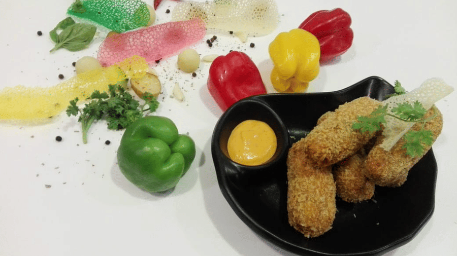 Black plate of breaded potato croquettes with a small bowl of orange dip, alongside whole bell peppers, herbs and tuile crisps on a white surface.