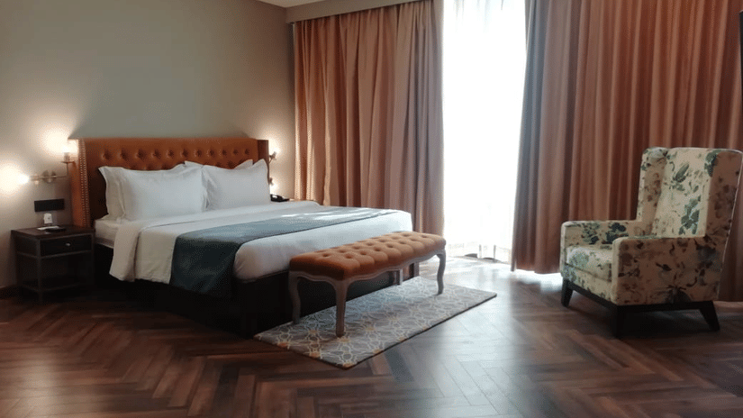 Suite bedroom with tufted tan headboard, upholstered bench, floral armchair and orange drapes over herringbone floor at Polo Hotel, Agartala.