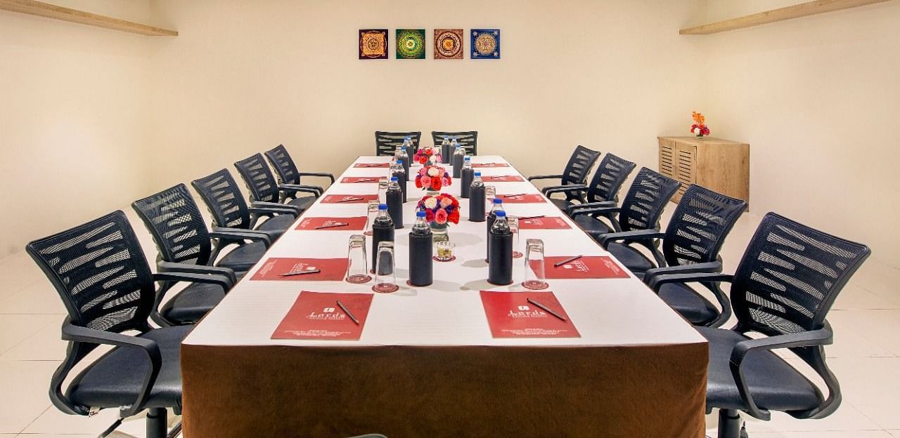 A formal meeting room is set up with a long rectangular table surrounded by numerous black office chairs, featuring water bottles and red and white placemats, under bright ceiling lights and with framed artwork on the wall - Lords Inn Motikhavdi, Jamnagar.