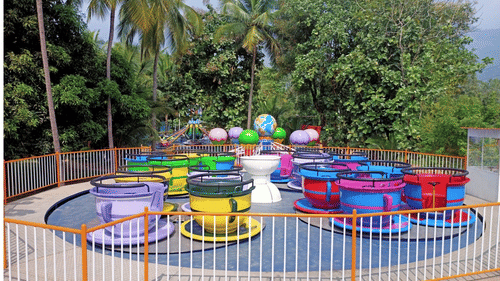A colourful amusement park ride with large teacup-shaped seats, set in a lush tropical environment with tall palm trees in the background.