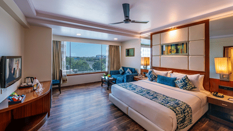 a king size bed room with a ceiling fan, a wall-mounted TV, a seating area, a work desk, and half length glass windows at Libra Lords Inn Jaipur