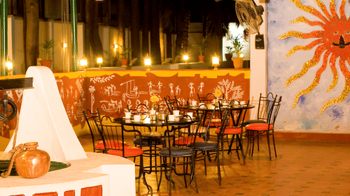 A restaurant area with tables, chairs, and a wall painting of a sun at the best resort in Bangalore | Ramanashree California Resort