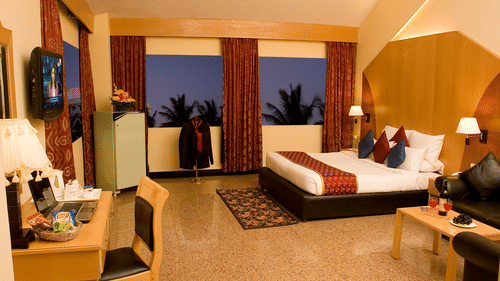 The Signet Suite room at the best resort in Bangalore, with a king sized bed, chairs, curtains, a wall mounted television and a table | Ramanashree California Resort