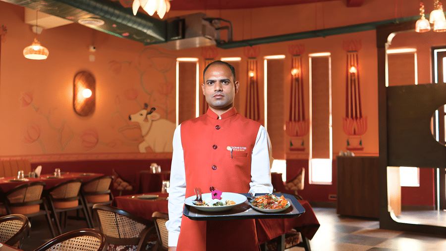 A waiter in Ekam restaurant at Kadamb Kuteer, Vrindavan wearing a red vest over a white shirt, holding a tray with food.