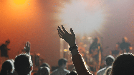 Silhouette of a crowd with hands raised at a live music concert, bathed in bright, atmospheric orange stage lighting.