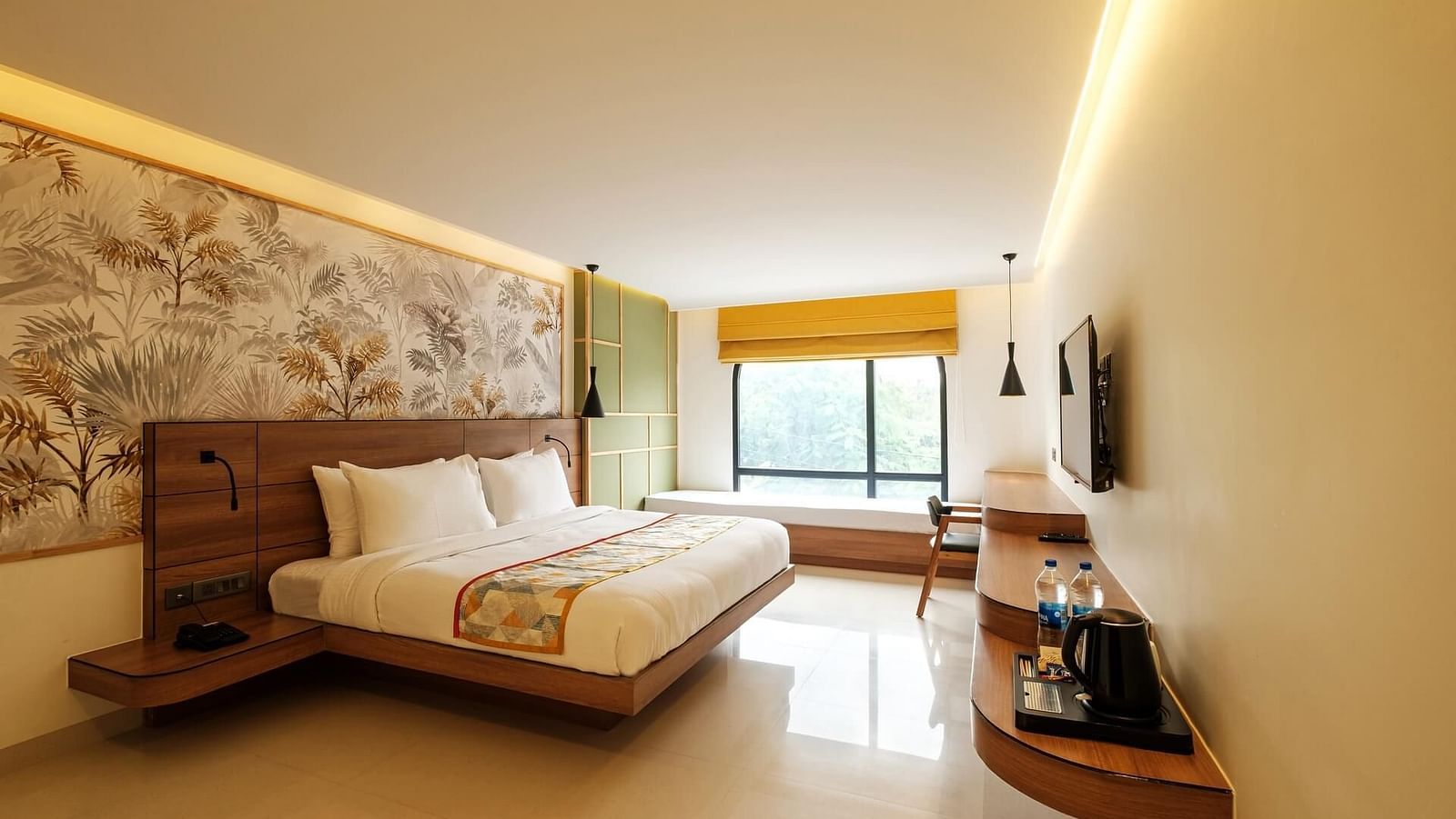 A spacious room at Indie Stays, Jaipur, with a large bed featuring a wooden headboard, a wall-mounted TV with a wooden shelf below, and a large window with an alcove.