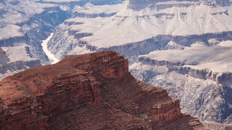 A high-angle view of a red rock plateau overlooking a winding river deep within a vast, layered canyon landscape.