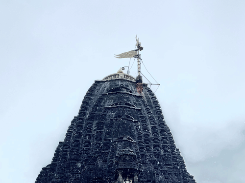 A tall stone temple tower viewed from below with a flag at the top against the sky with intricate art on the exterior.
