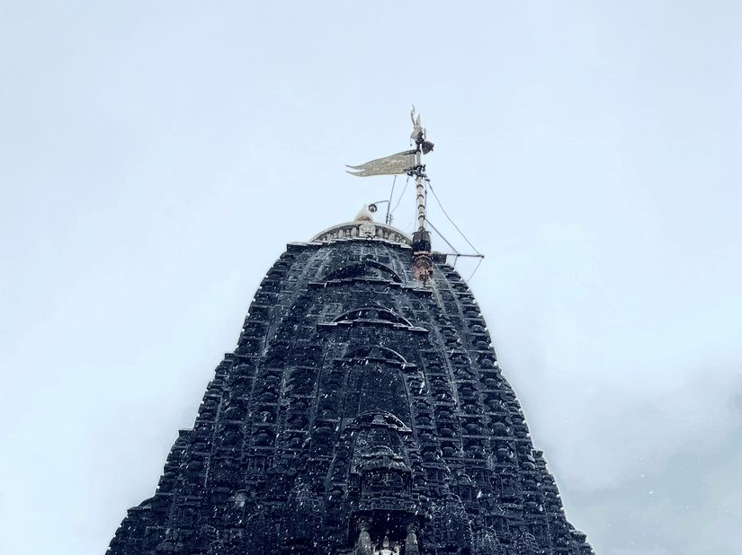 A tall stone temple tower viewed from below with a flag at the top against the sky with intricate art on the exterior.