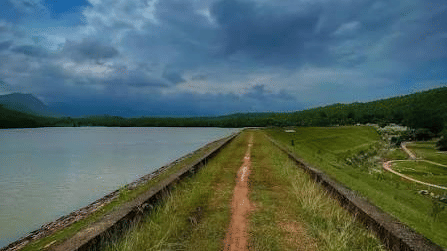 A path running along a body of water of Sankarmara Dam under a cloudy sky with hills in the distance near Khairi Resorts.