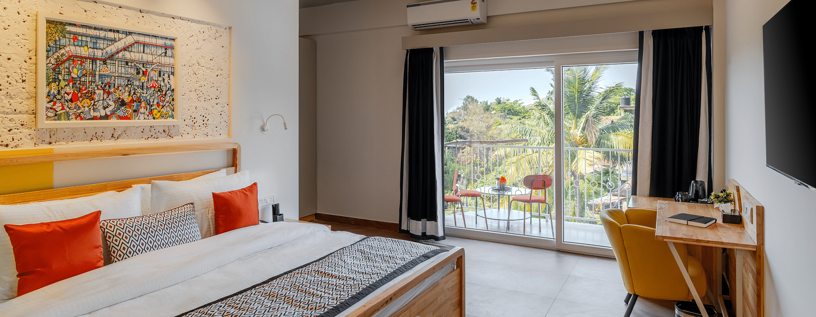 Bright hotel room with, flat TV screen, work desk and chair, vibrant art on the wall, and an access to the balcony at Bedzzz Xclusiv Morjim, Goa