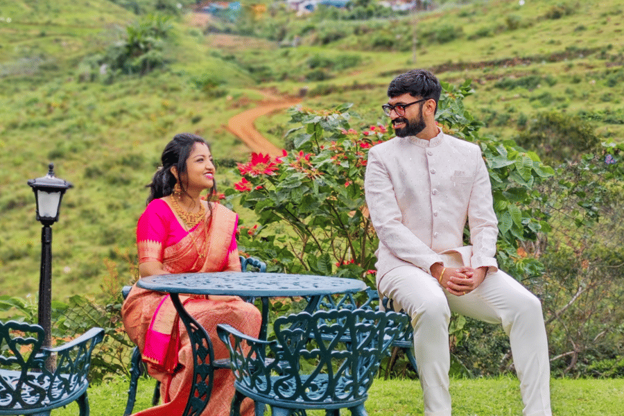 A couple is seated at a small metal table on a grassy lawn with a scenic hilly background; the woman wears a pink sari, and the man wears a white suit.