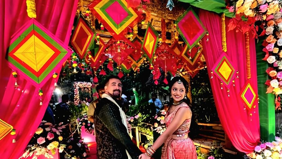 A decorated outdoor wedding venue entrance features colorful fabrics, floral arrangements, and traditional Indian elements, with people gathered around