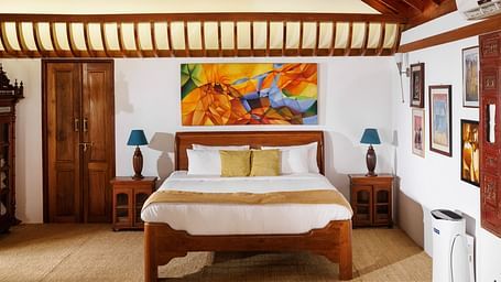A room with wooden interiors featuring a bed, bedside tables, and paintings hung on the wall - BluSalzz Villas - Muhamma Resort, Alleppey