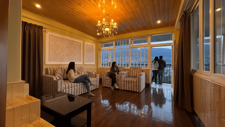 A couple sitting on a hotel room balcony at night, enjoying a peaceful and romantic view of the lit-up Kurseong city and the Giddha Pahar mountains.