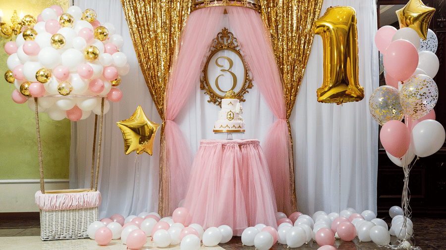 Decorated first birthday setup with a child standing in front of a pink backdrop, balloons, and a golden number 1 balloon.