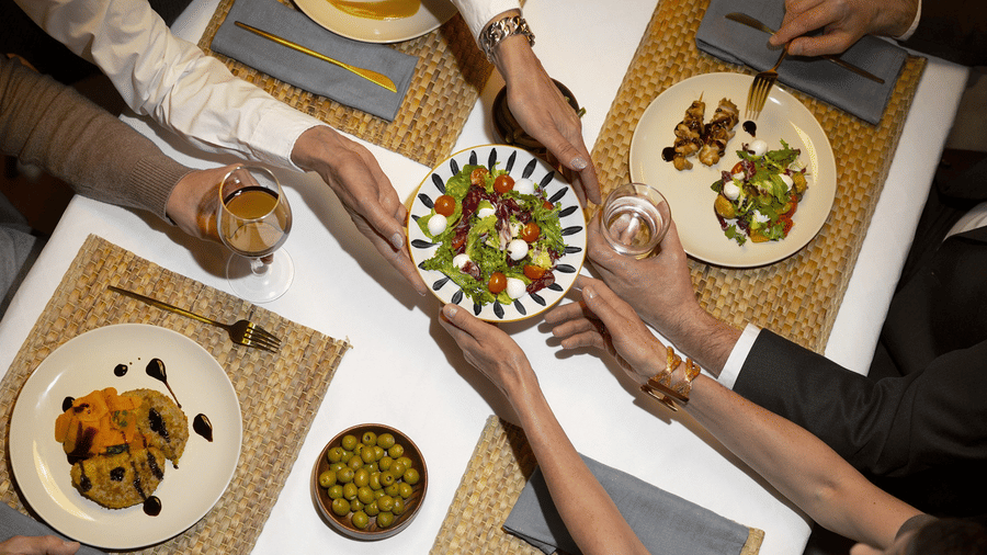 Group of people sharing a meal at a table, with hands reaching for food, emphasizing communal dining and interaction.