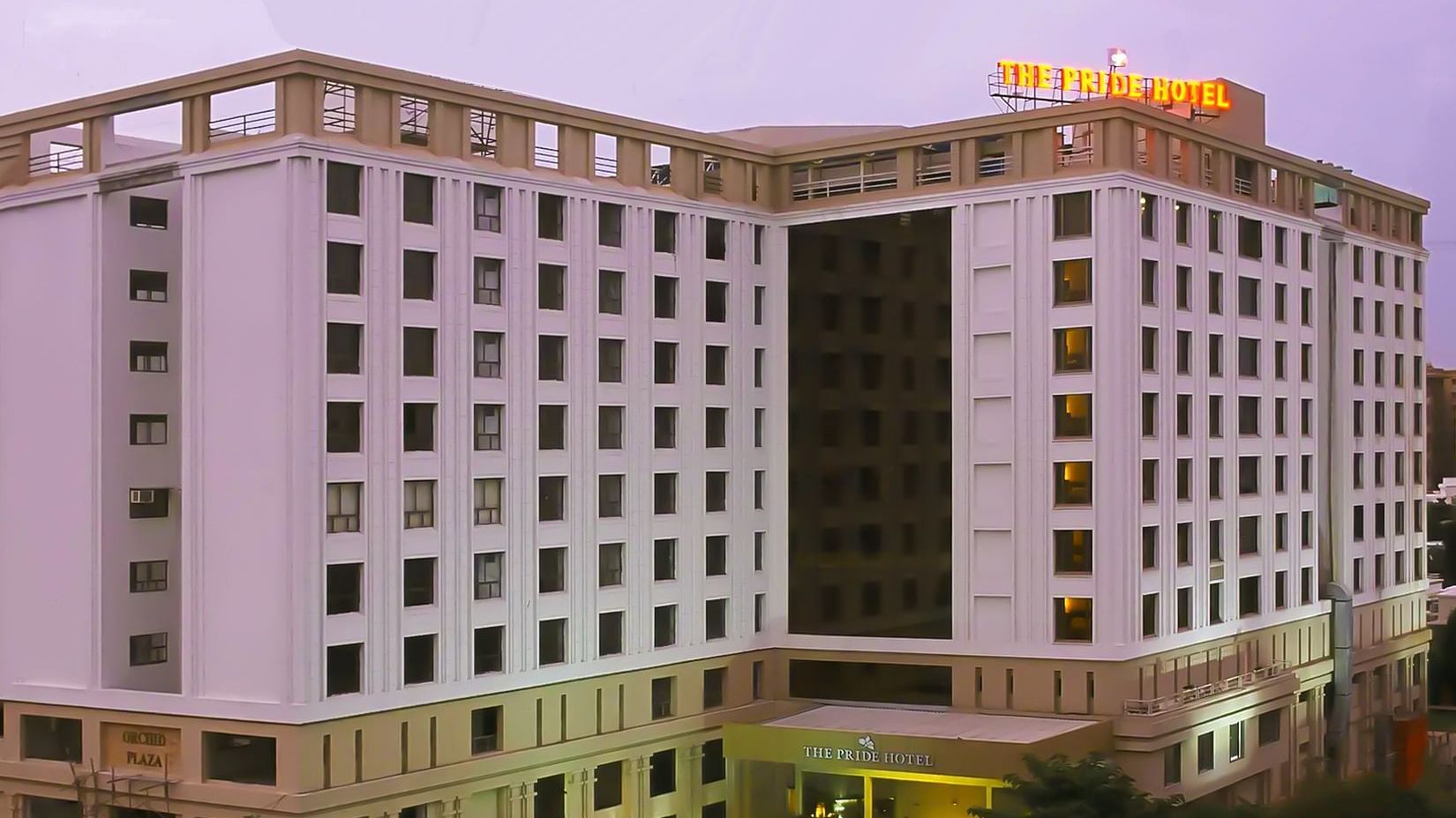 Facade of Pride Plaza Ahmedabad hotel with its name lit up atop the building, set against a glowing sunset sky for a striking evening view