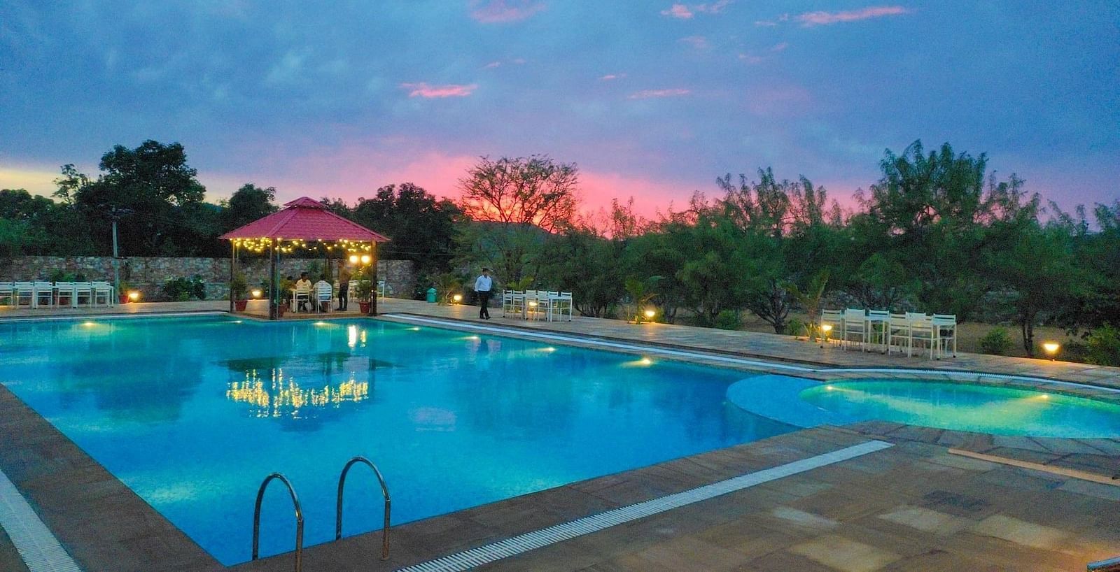 An outdoor swimming pool at dusk with a pink and blue sky in the background - Bamboo Saa Mulberry Resort, Pushkar