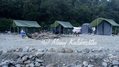 Green camping tents set up on 1 rocky riverbank backed by a lush forest hillside.