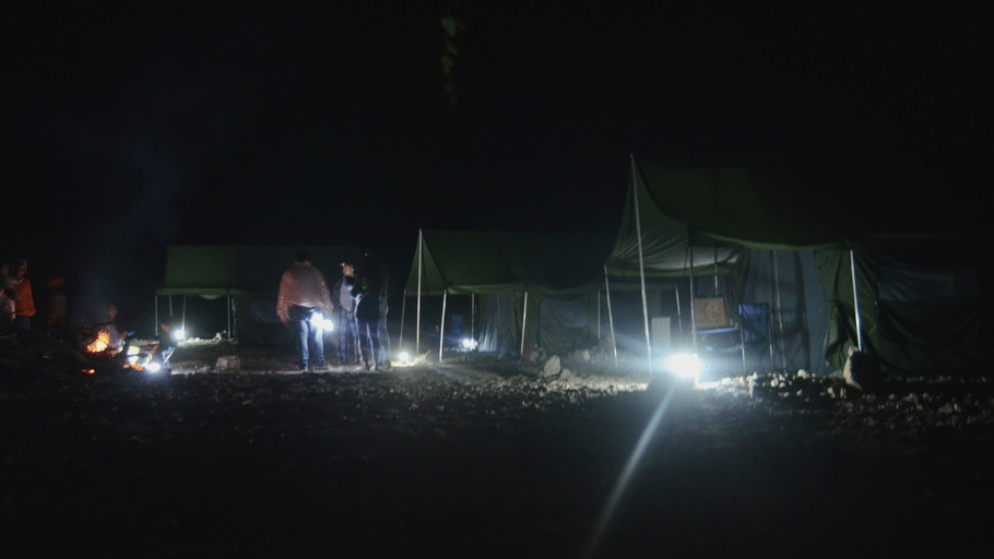 A campsite at night featuring illuminated tents, a glowing campfire, and bright lanterns under a dark sky.