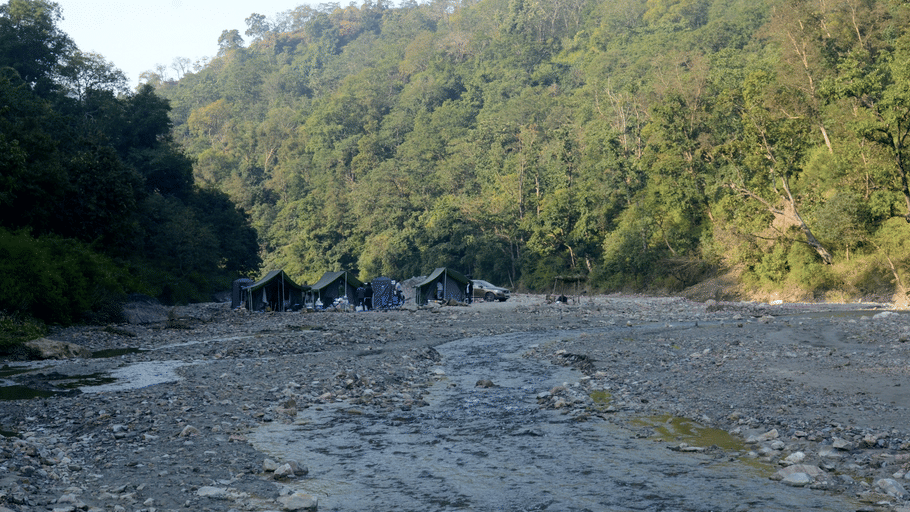 A dry rocky riverbed with small streams surrounded by forested hills and a distant campsite.