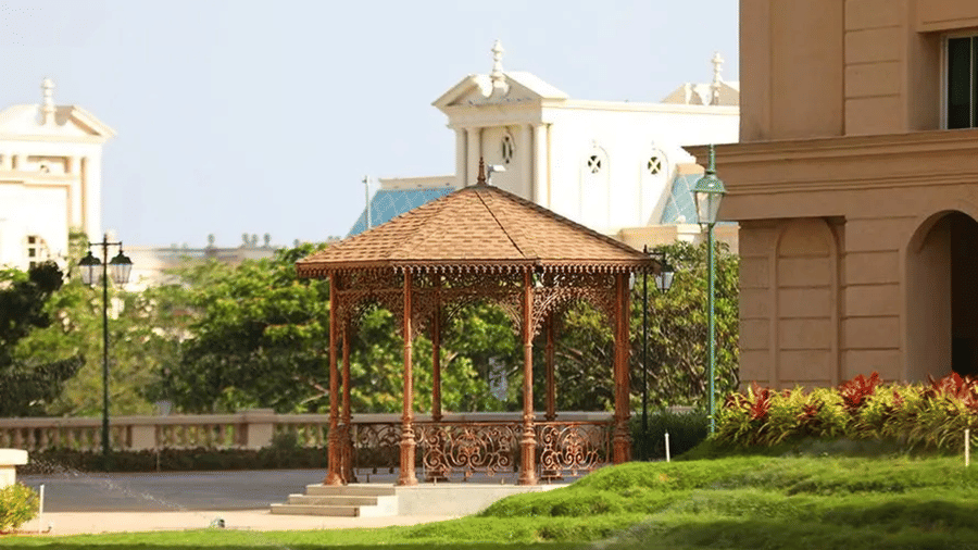 A decorative wooden gazebo stands on a manicured green lawn within a luxury housing estate, featuring elegant, classical-style architecture behind at Apartel by Aarin, Oragadam.