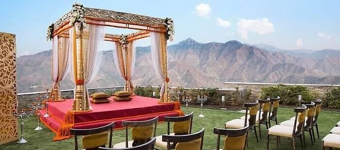 a mandap set up meant for wedding ceremony on manicured lawn with chairs facing it and in the background there are hils at a distance- neer ganga Resorts