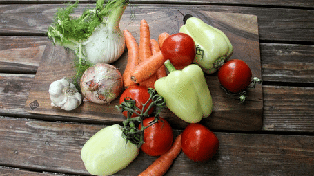 An assortment of fresh raw vegetables, including tomatoes, peppers, carrots, fennel, and garlic on a wooden table.