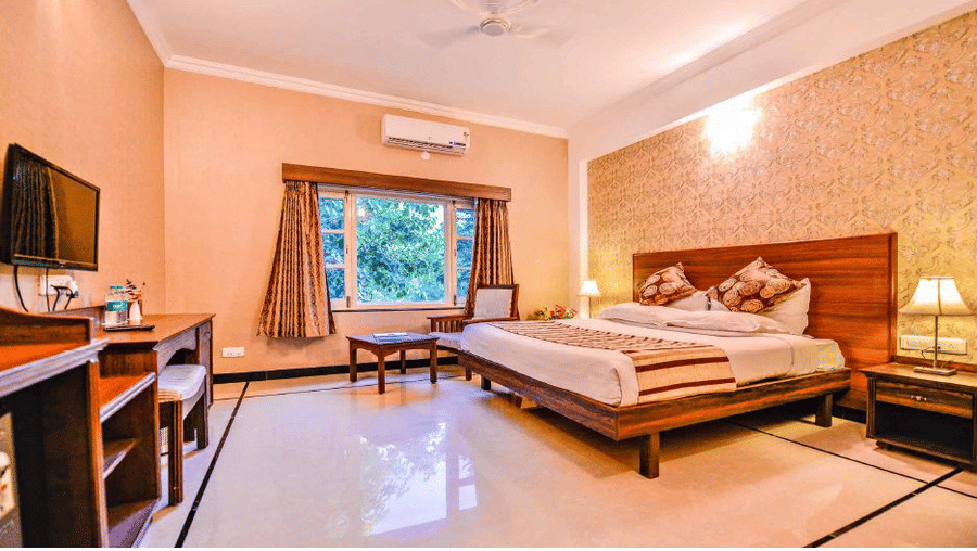 A bedroom with a king size bed, a writing desk, a wall-mounted television, and a window at Amantra Shilpi Resort, Udaipur.