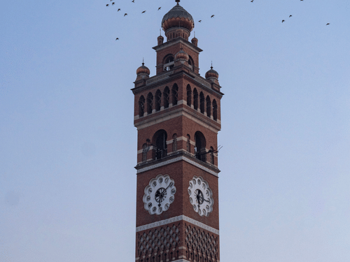 The Hussainabad Clock Tower in Lucknow standing tall against a clear blue sky in the background, with a flock of birds flying around the tip of the tower.